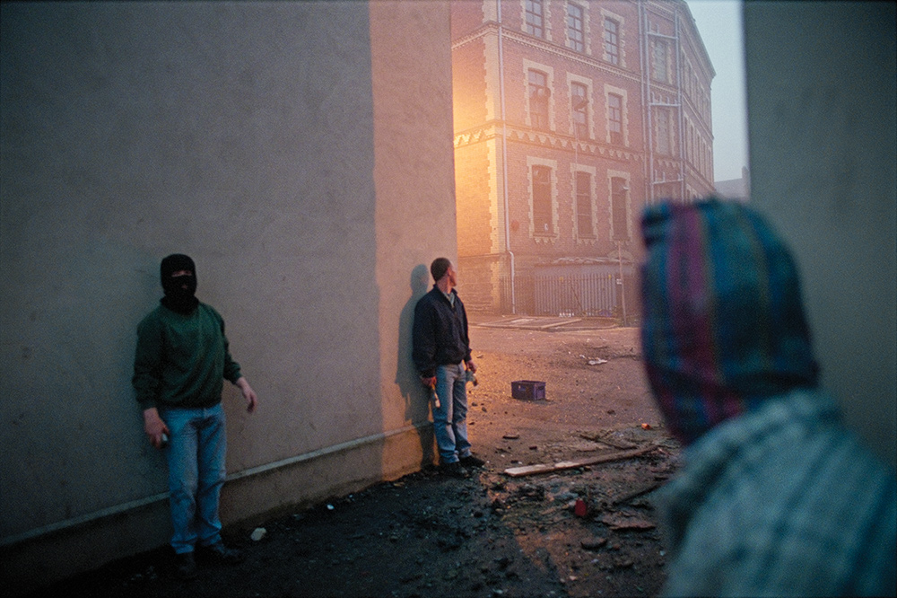 Three men with masks or obscured faces stand along an alley wall, waiting. There is rubble in the street.